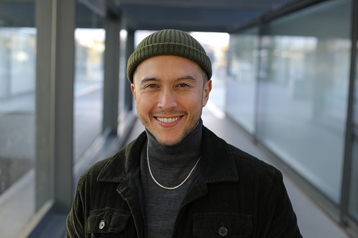 A very cute looking Hispanic man is posing with a gorgeous smile. He wears a modern outfit with a beanie and necklace. His style is hip and fresh.