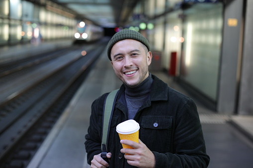 A very handsome Hispanic young man is waiting for a train. He holds a coffee cup and smiles.