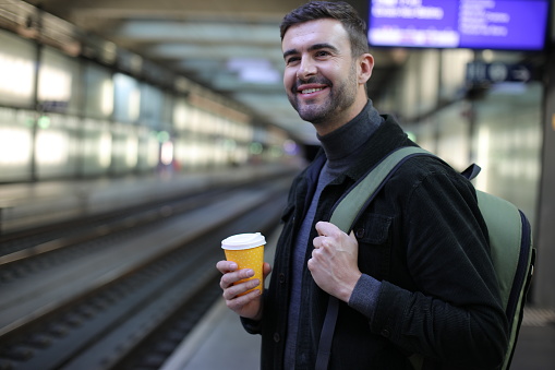 A very handsome man is waiting for a train. He holds a coffee cup and smiles.