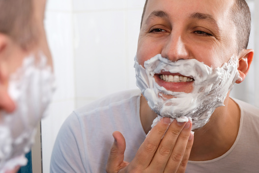 Happy man shaving in front of a mirror in the bathroom. Hygiene, skin care. The guy shaves his beard and mustache.