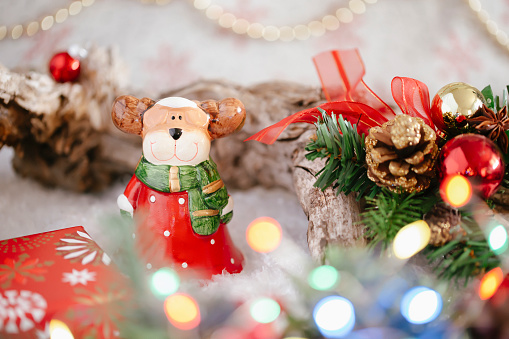 Close-up Christmas holiday background with a ceramic elk dressed as Santa Claus among the Christmas tree with cones, balls, bokeh, copy space.
