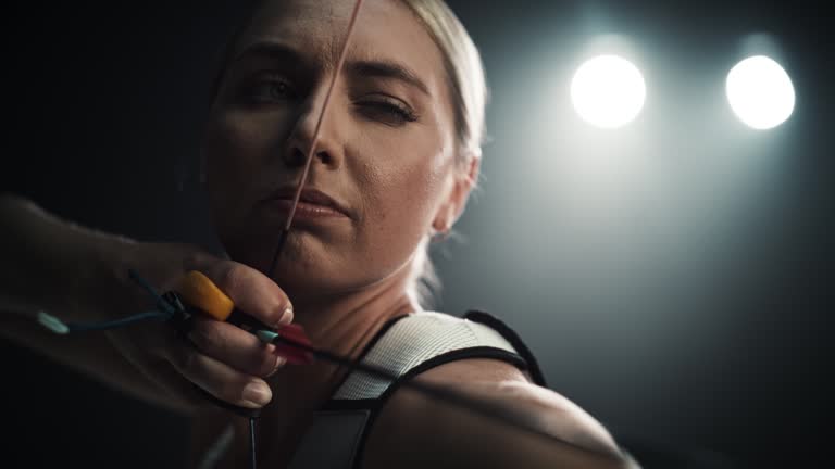 Cinematic Handheld Footage of a Professional Female Archer in Action. Portrait of a Beautiful Athlete Holding a Bow and Pulling Back the Arrow in Order to Hit the Target in a Dark Competition Room