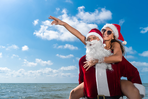 Portrait of a happy Santa Claus holding on his back a smiling young woman in a Santa hat with a pointing finger to the side on the seashore. Celebrating Christmas holidays in warm countries