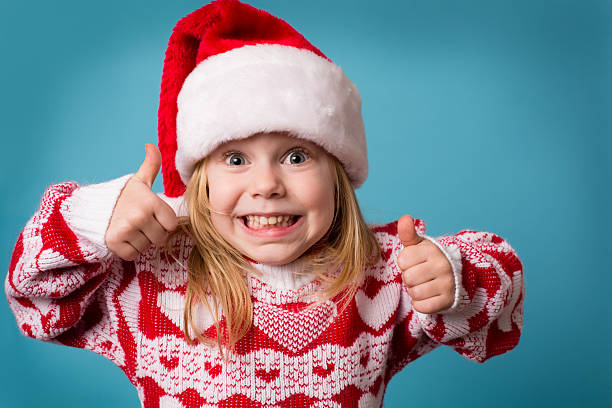 Santa's Little Helper Giving Thumbs Up Sign "Color image of a little girl wearing a Santa hat and an ugly Christmas sweater, smiling and making the thumbs up sign." christmas excitement stock pictures, royalty-free photos & images