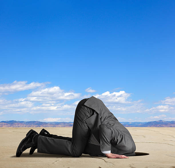 Businessman burying his head in the sand A businessman, on the ground and on all fours, buries his head in a hole. The scene is set in a large desert like environment.  A range of mountains can be seen on the horizon. Clouds hang over the mountain range and the blue sky above leaves ample room for text. Image can be easily cropped horizontal or vertical. head in the sand stock pictures, royalty-free photos & images