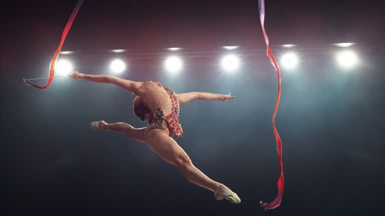 Graceful Rhythmic Gymnast Performs a Powerful Split Leap with a Ribbon. Young Female Exercising in a Striking Beautiful Uniform in a Dark Room. Cinematic Super Slow Motion Speed Ramp Footage