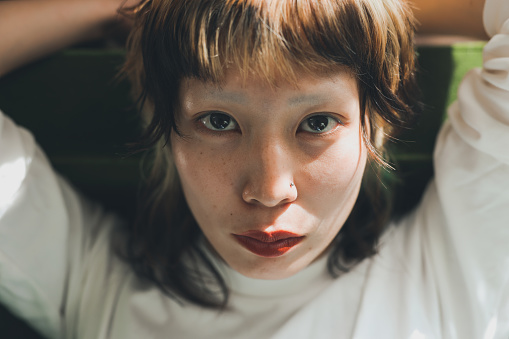 Close up shot of young asian Gen-Z female with colorful hair looking at camera on the sofa at home under the sun cast through the window.