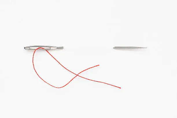 A sewing needle piercing the white background. A short piece of red thread is threaded through the eyelet. Isolated on white.