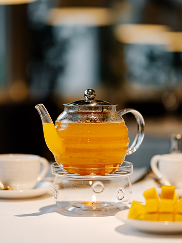 Herbal tea with mango fruit in transparent glass teapot with warmer and candle on table in restaurant. Herbal tea concept, wellbeing and health care. Vertical.