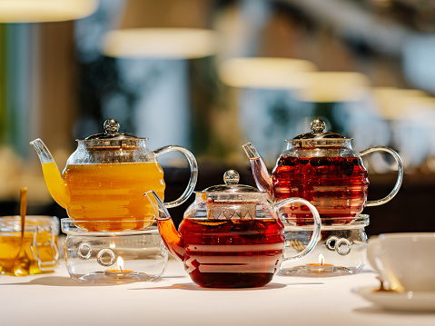 Set of three transparent glass teapots with warmer and candles, hot herbal tea on table on restaurant bokeh background. Mango tea, rosehip herbal tea, spicy herbal tea with star anise and honey - tea concept