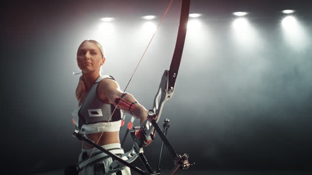 Slow Motion Speed Ramp Portrait Footage of a Female Archer Aiming and Shooting at a Target with a Modern Compound Bow. Young Athlete Drawing and Releasing the Arrow in a Dark Studio for Advertising