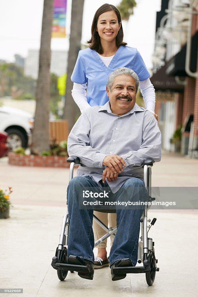Carer Pushing Disabled Senior Man In Wheelchair Carer Pushing Disabled Senior Man In Wheelchair smiling To Camera. Latin American and Hispanic Ethnicity Stock Photo