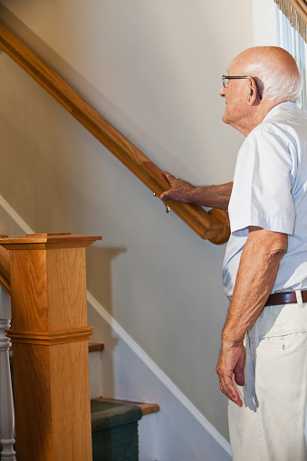 Elderly man (80s) standing at bottom of staircase, looking up.