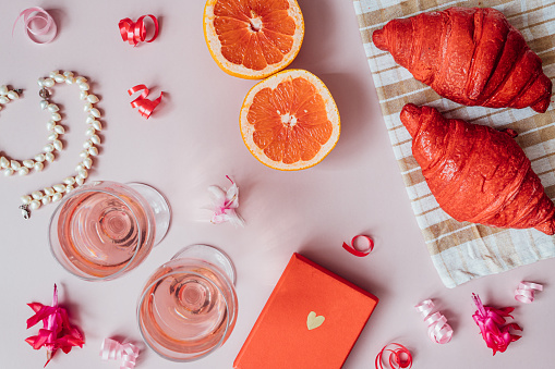 top view, close-up, a festive romantic breakfast on valentine's day on a pink background, a red croissant, a red box with a gift, two glasses of rose wine or champagne, pearl necklace
