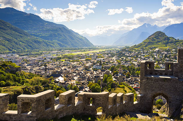 Panoramic landscape of Sion town nestled in Swiss Alps under clear blue sky, showing valley, historic buildings and mountainous terrain.