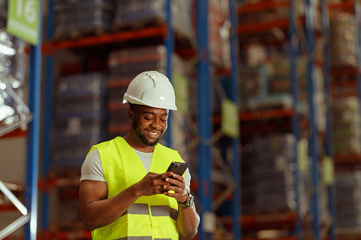 A Black Warehouse worker is using a Smartphone in the Warehouse. He is wearing a reflective vest and a protective helmet.