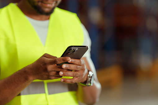 A Black Warehouse worker is using a Smartphone in the Warehouse. He is wearing a reflective vest and a protective helmet.