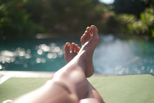 Close-up POV shot of unrecognizable woman legs crossed at ankle lying next to poolside. She's sunbathing in a hotel