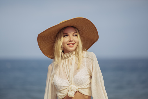 Stunning blonde woman in summer beach outfit relaxing outdoors against sea background. A fashionable romantic young adult lady wearing a trendy vintage straw hat, white blouse, and skirt, standing at the beach