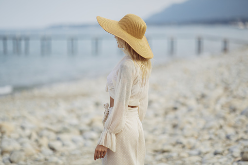 Stunning blonde woman in summer beach outfit relaxing outdoors against sea background. Back view of fashionable romantic young adult lady wearing a trendy vintage straw hat, white blouse, and skirt, standing at the beach