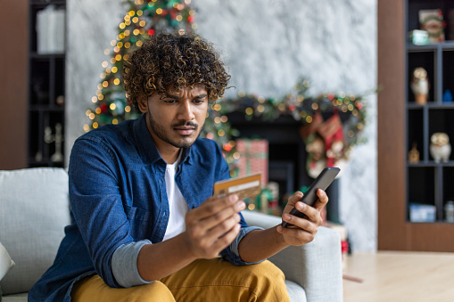 Cheated and upset Christmas man in living room near decorated Christmas tree, rejected and wrong money transfer, hispanic man holding bank credit card and phone.