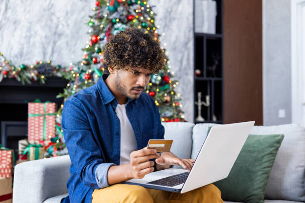 istock Man trying to make a purchase in online store, received a rejection, error transfer of funds, hispanic on Christmas, sitting near the tree in the living room with laptop, holding a bank credit card 1752304721