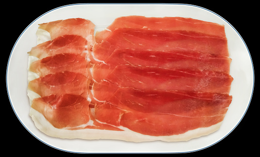 Slices of Prosciutto offered on oval blue rimmed white oval porcelain tray, isolated on black background, high resolution stock image.