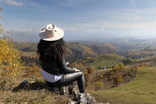 Young woman sitting on a rock wall and admiring the beautiful autumn landscape in front of her eyes.