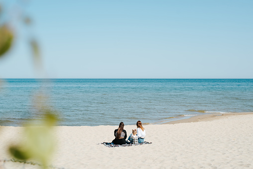 Back view of a couple, woman and little girl, relaxing together on the beach by the sea, sitting on a blanket outdoors on a sunny day, general view.