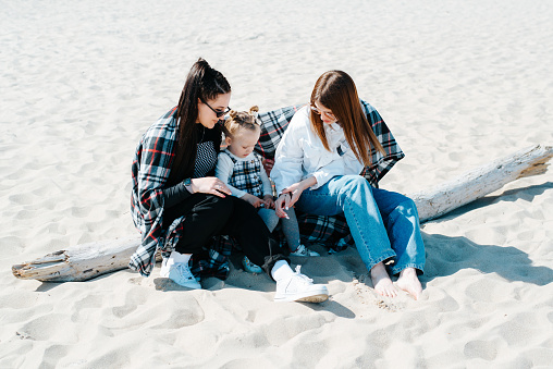 Couple of women girlfriends and little daughter spending time together on beach sitting on a log.