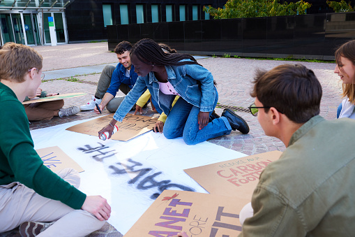 Multiracial group of student protestors making placards and banners with spray paint on city street for climate change