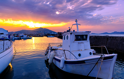 Colorful sunset over quiet Greek fishing village and its harbour, old traditional boat moored by pier, sun rays and clouds in dramatic sky. Distant hills and hazy mountains, beautiful landscape or seascape of Naoussa, Paros island, Greece.
