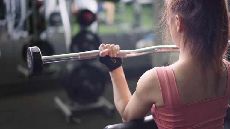 Back view of beautiful Asian lady executing a barbell curl in the gym