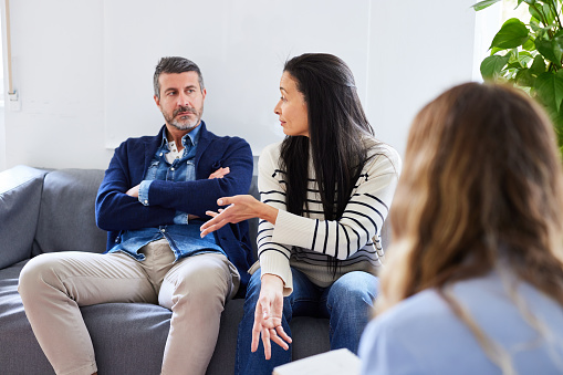 Anger couple sitting on sofa and arguing with each other in front of psychotherapist during marriage counseling session in office