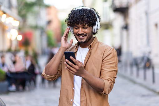 Young Indian man standing on city street wearing headphones and phone and enjoying music, smiling.