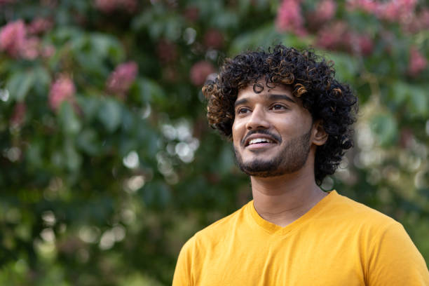 nahaufnahme. porträt eines jungen indischen mannes, der in einem orangefarbenen t-shirt draußen in einem park steht und lächelnd zur seite schaut - hinduism outdoors horizontal close up stock-fotos und bilder