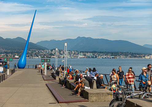 Vancouver, Canada - 10th September 2023: visitors mix with locals strolling or sitting on seats on the Harbourfront walkway, Coal Harbour, on a sunny September day. The blue sculpture is The Drop, designed by Inges Idee and installed in 2009. It is an homage to the power of nature, and locally regarded as an 'in joke' referring to Vancouver's high rainfall.