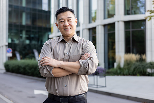 Portrait of a young Asian man standing near an office center, with his arms crossed on his shoulders and looking at the camera, with a smile.