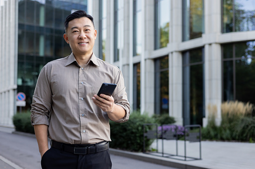 Portrait of a young Asian male lawyer standing near an office building, holding his hand in his pocket and using the phone. Smiling and looking at the camera.