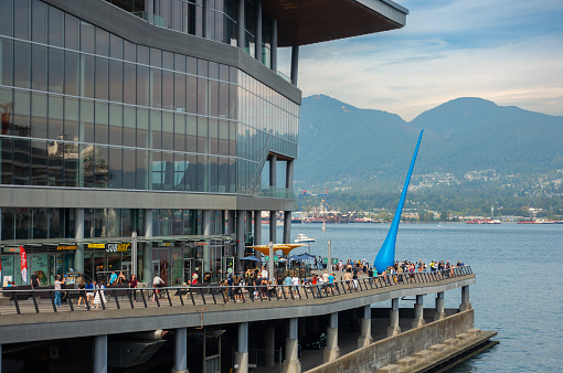 Vancouver, Canada - 10th September 2023: visitors mix with locals on the Harbourfront walkway in front of (or behind!) Vancouver Convention Centre. The blue sculpture is The Drop, designed by Inges Idee and installed in 2009. It is an homage to the power of nature, and locally regarded as an 'in joke' referring to Vancouver's high rainfall. North Vancouver and Grouse Moiuntain are in the background.