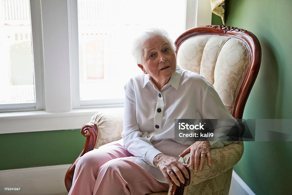 Senior woman sitting in chair by window Senior woman (80s) at home sitting in chair by window. Corner Stock Photo