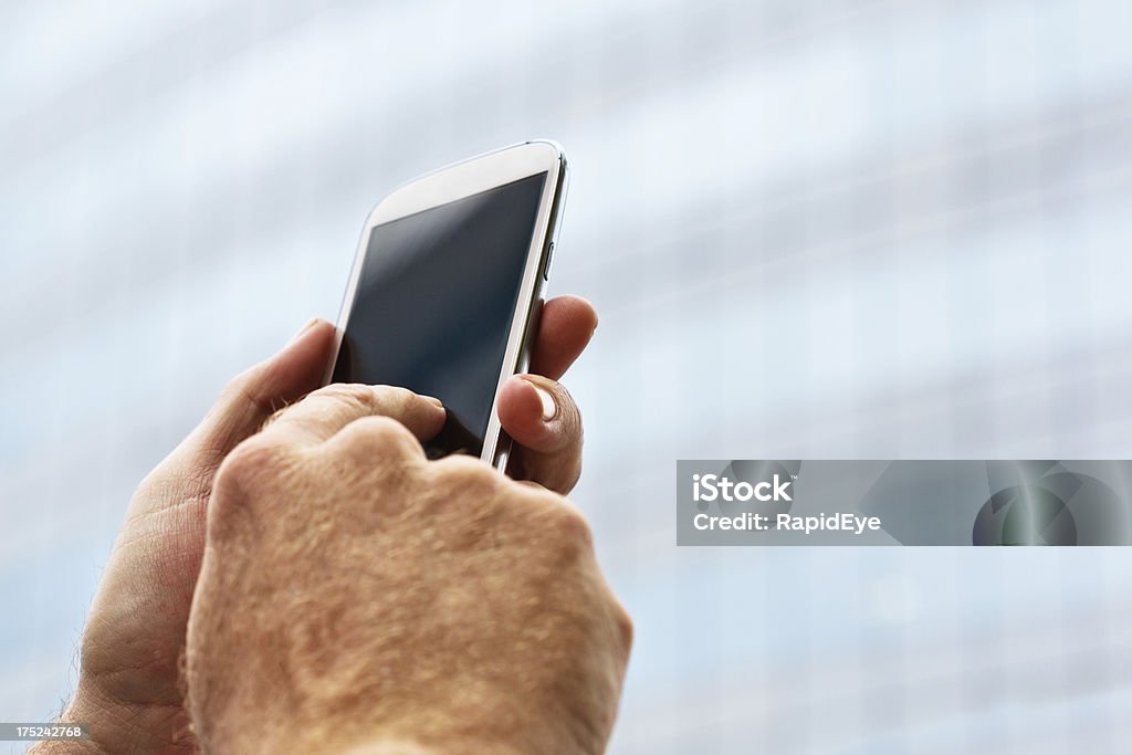 Searching for a signal, male hand holds up smart phone "A man holds a smart phone aloft, perhaps searching for a signal, tapping the blank screen. The exterior of an out-of-focus office building is in the background." Blank Stock Photo