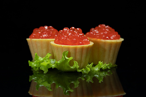 Three tartlets stuffed with red salmon caviar and decorated with lettuce leaf. Tasty snack for Christmas or New Year dinner. Black background.