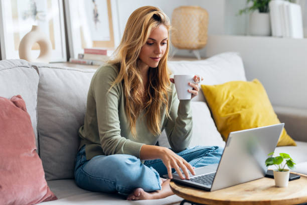 Confident young woman working with her laptop while drinking a cup of coffee sitting on a couch at home