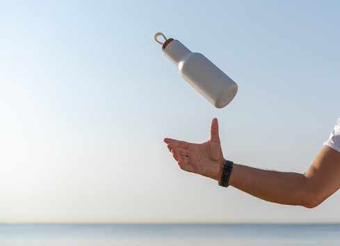 Hand throwing a modern metal water bottle, sea surface on the background