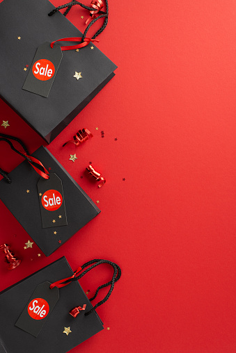Unbeatable Black Friday bargains await! A top-view vertical shot of discount labels, paper bags, festive tinsel, and starry confetti on a red background, leaving space for your promo text