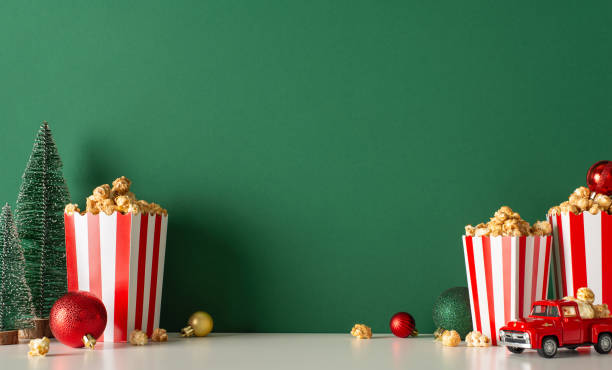 istock Enjoy cozy Noel celebration at home with popcorn delivery theme. Side-view image of table featuring tiny red car, popcorn in boxes, baubles, small fir against green wall backdrop, room for movie promo 1752469486