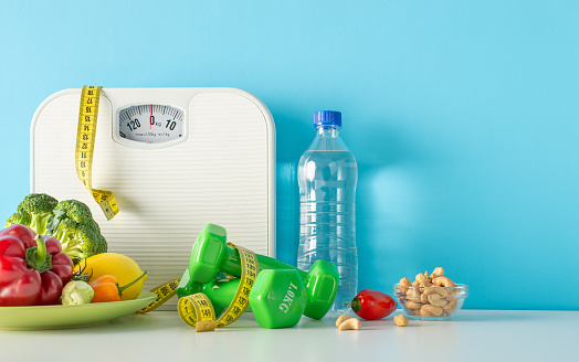 Nutrition and fitness concept: Table side view setup with scale, measuring tape, dumbbells, nuts, and a variety of fruits and vegetables on blue wall background, leaving space for text or advertising