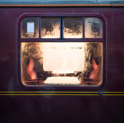 Looking through the glowing window of an old fashioned train carriage at tables and seats, with water condensation on the window.