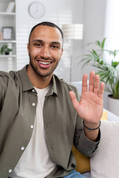 istock Vertical photo. A young smiling Muslim man is doing an online video broadcast from home. Sitting on the couch and waving at the camera 1752476476